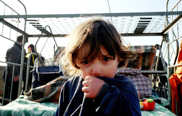 A young child sits on a bed, facing the camera, with a backdrop of refugees.